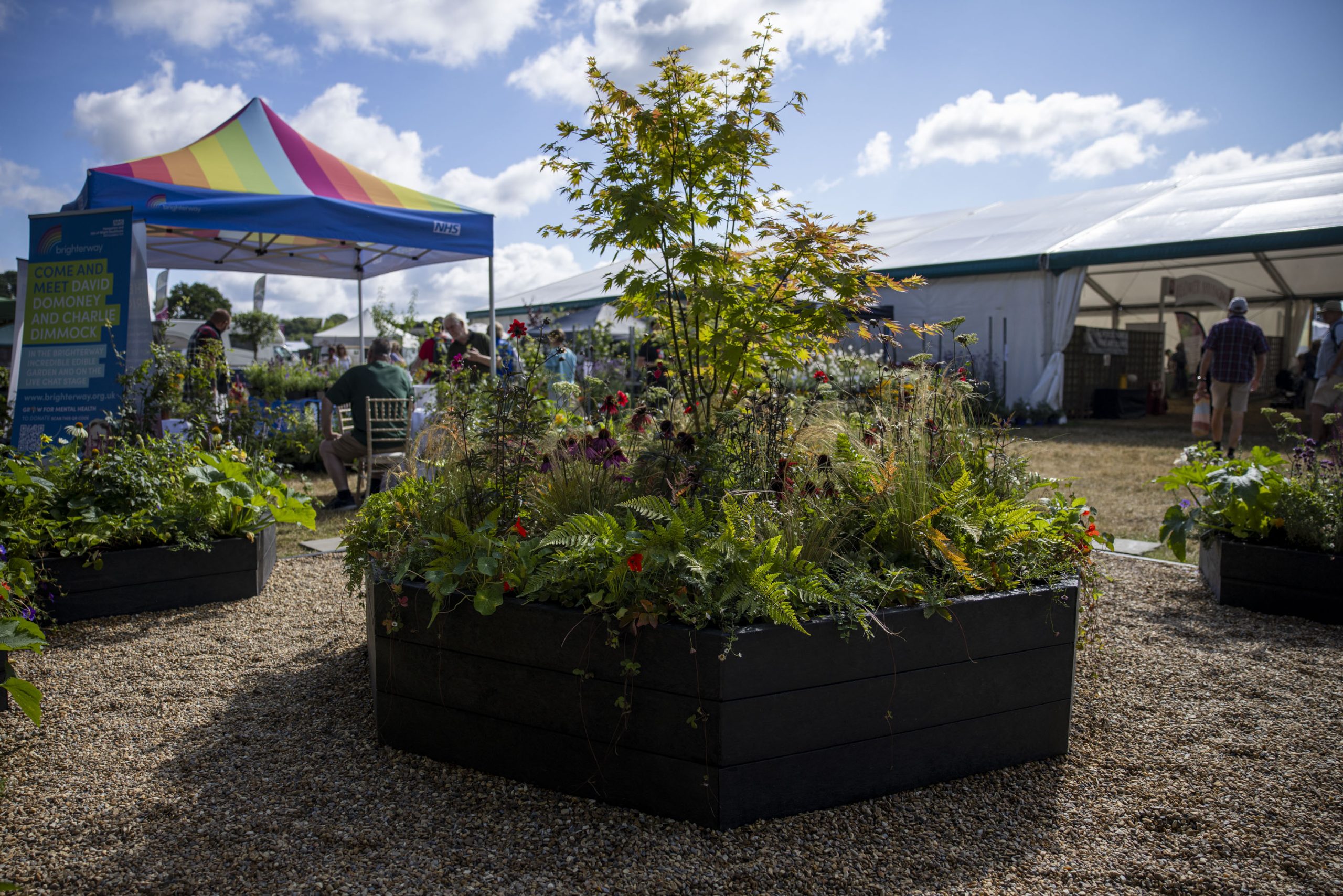 A bright tent to frame left with a garden show display in the centre, with blue sky and other garden show exhibits in the background