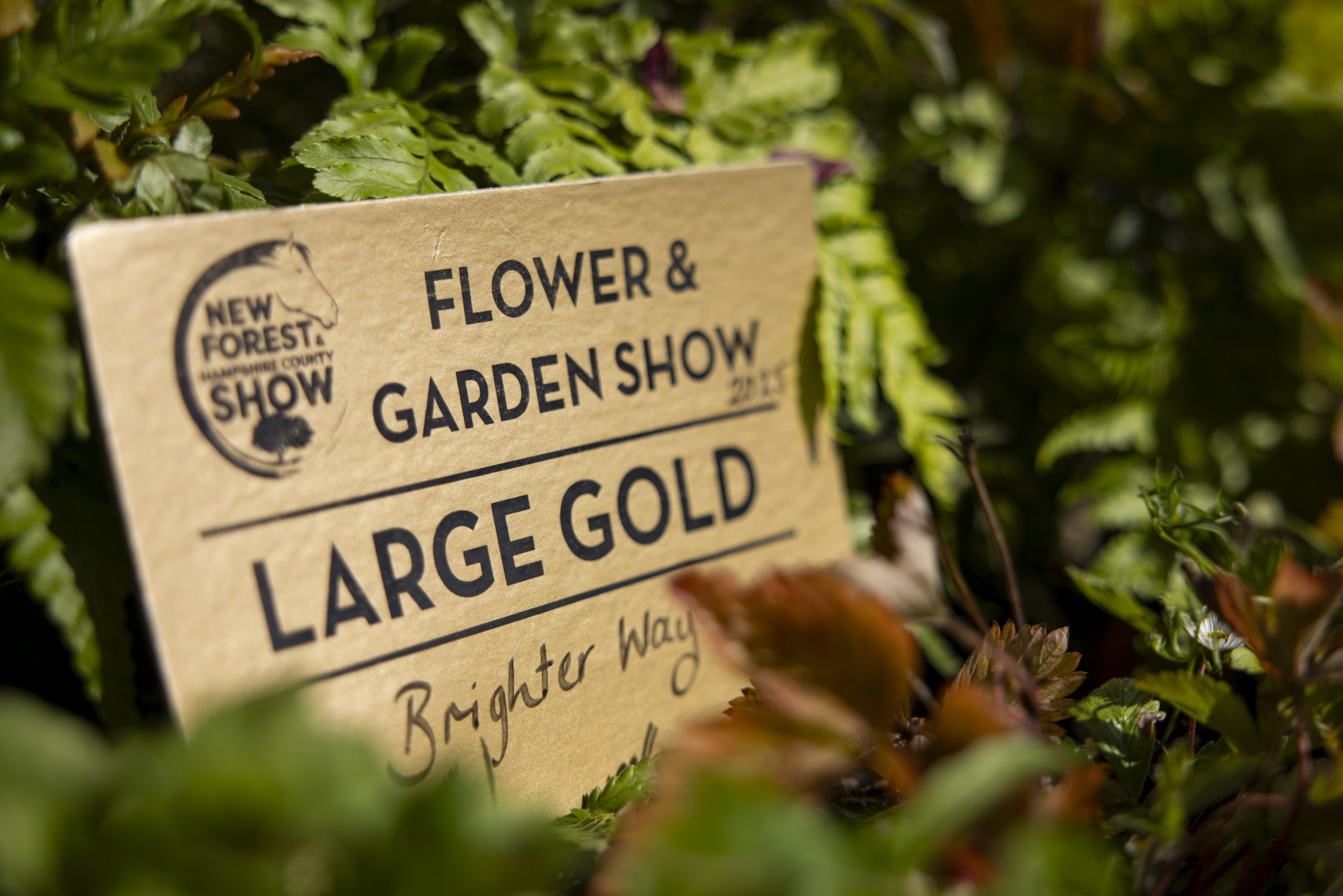 A sign framed by lush plants reading 'Flower & Garden Show - Large Gold - Brighterway'
