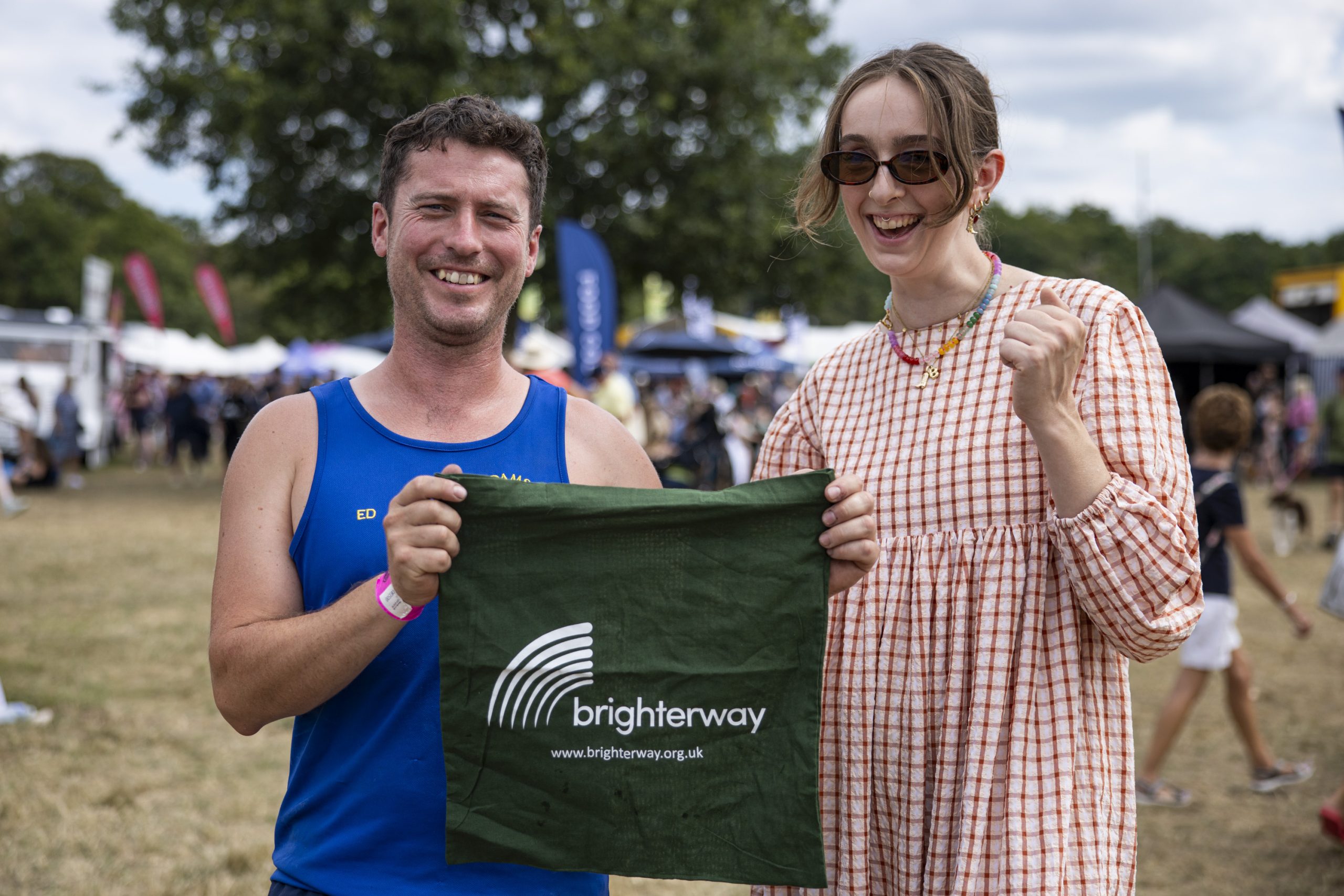 A man in a blue vest and a woman in a checkered frock and sunglasses hold up a green Brighterway t-shirt between them