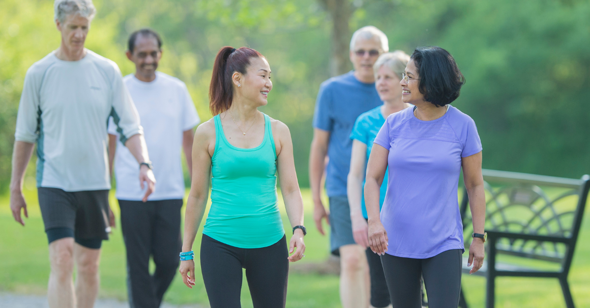 Six middle aged men and women walk in a group, smiling and talking, dressed in comfortable walking and running attire.