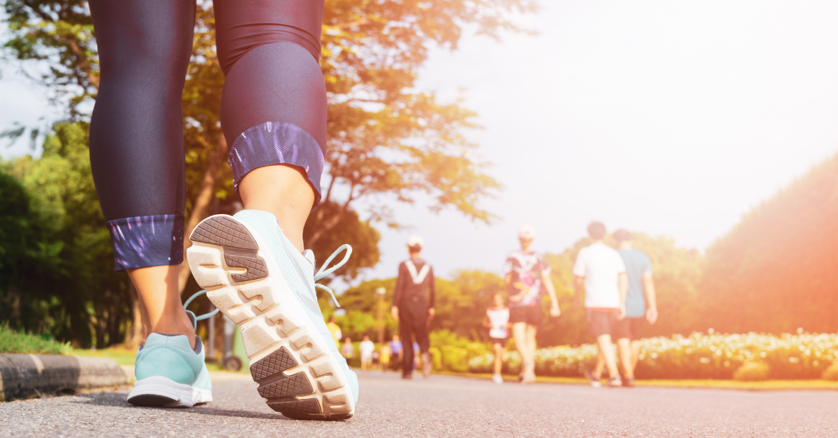 Close up of a woman's lower legs in running tights and running shoes, walking away from the camera on a tarmac path. Other people appear in the background, which features trees and grass. The image has a warm lens flare effect to frame right.