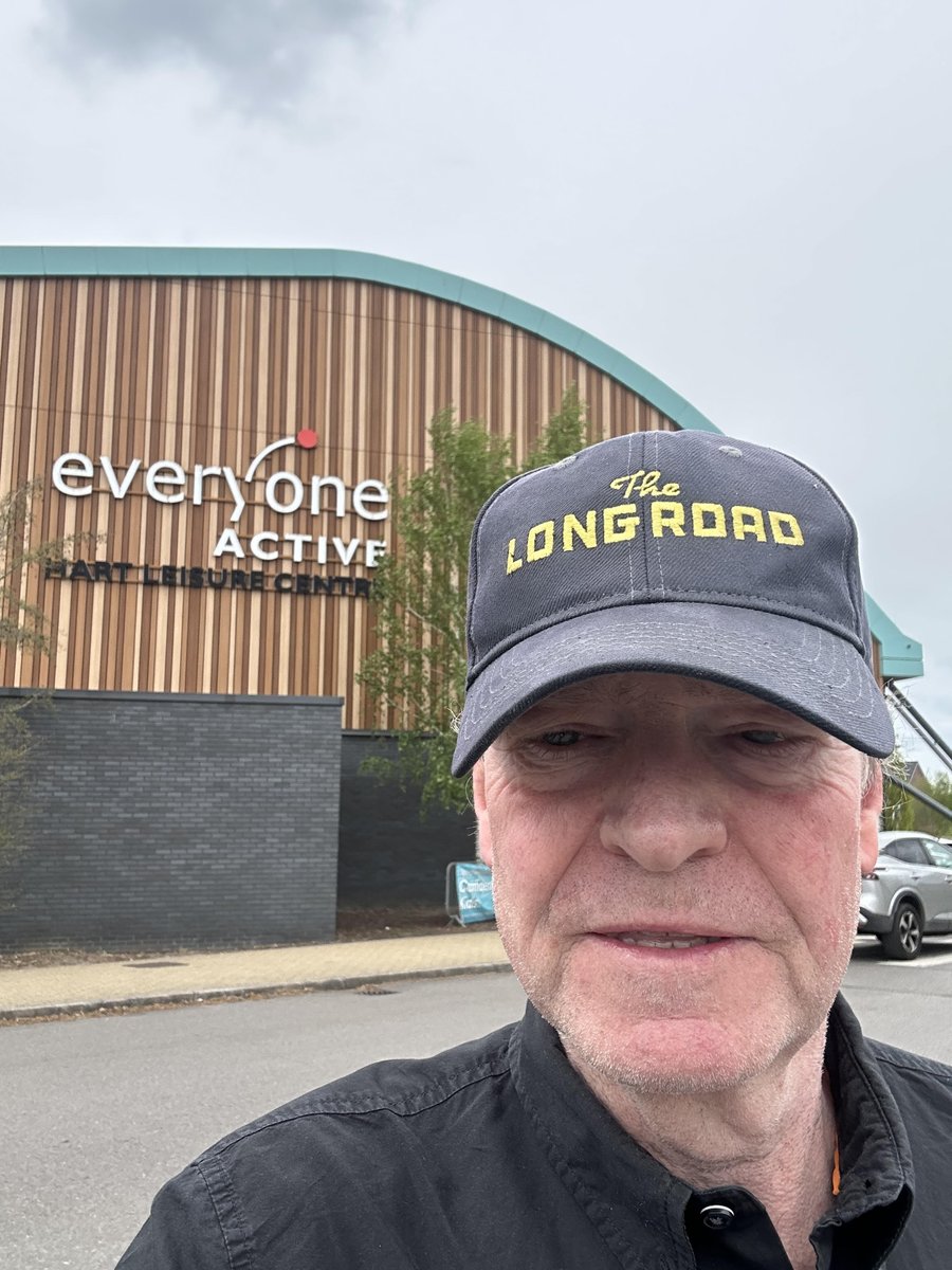 A middle aged man in a cap stating 'The Long Road' stands in front of an Everyone Active gym with a wood facade and car park behind the man.
