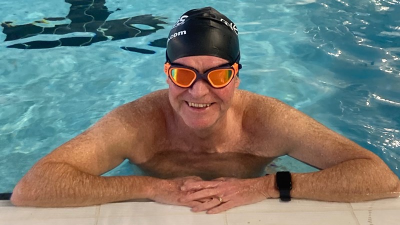 A smiling man in swimming goggles and swimming cap stands in a swimming pool, resting his arms on the edge.